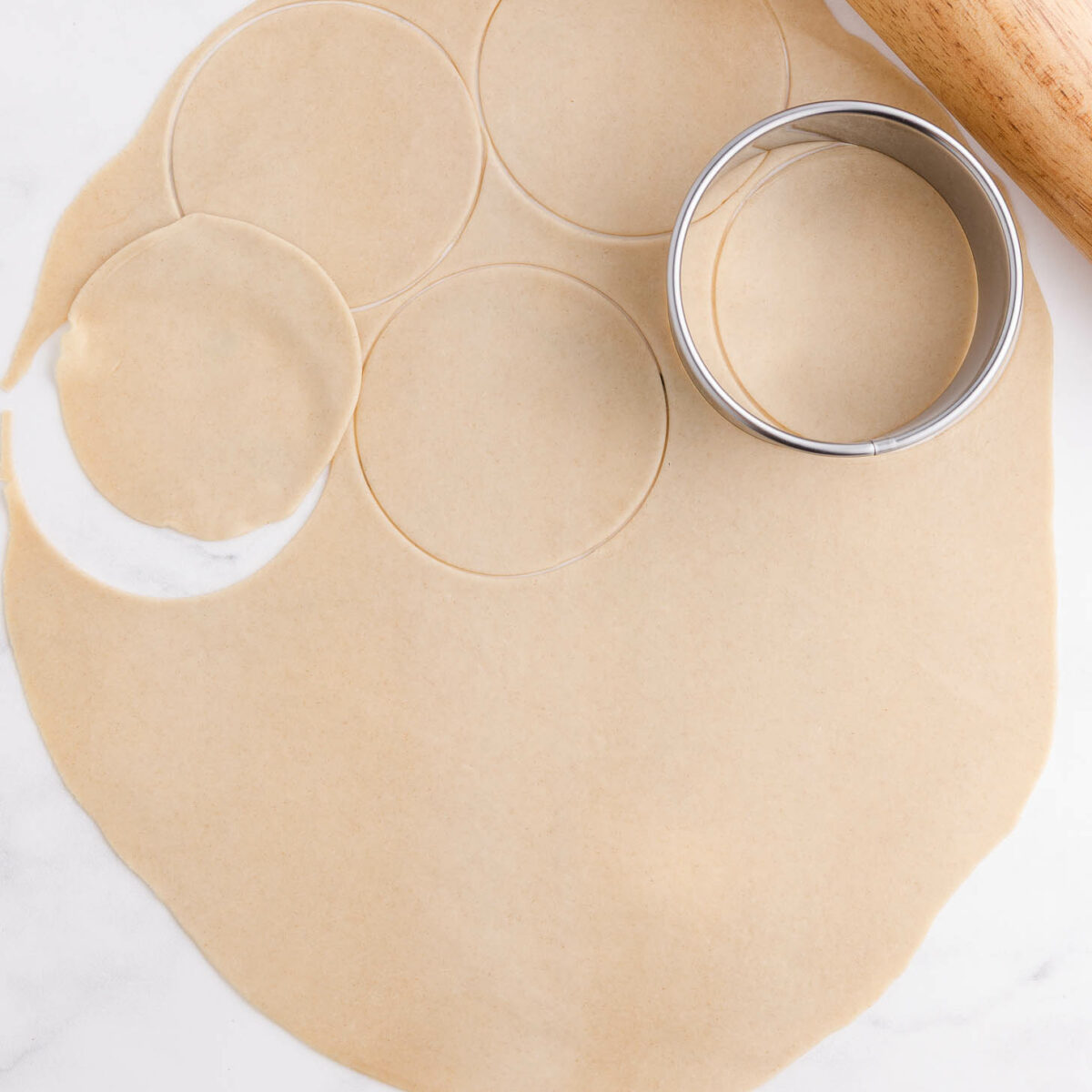 A rolled-out sheet of dough with circular shapes cut out, a round metal cutter, and a wooden rolling pin on a white surface.
