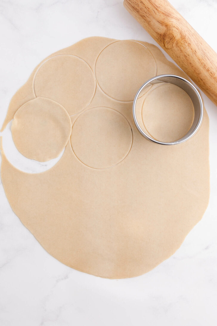 A rolled-out sheet of dough with circular shapes cut out, a round metal cutter, and a wooden rolling pin on a white surface.