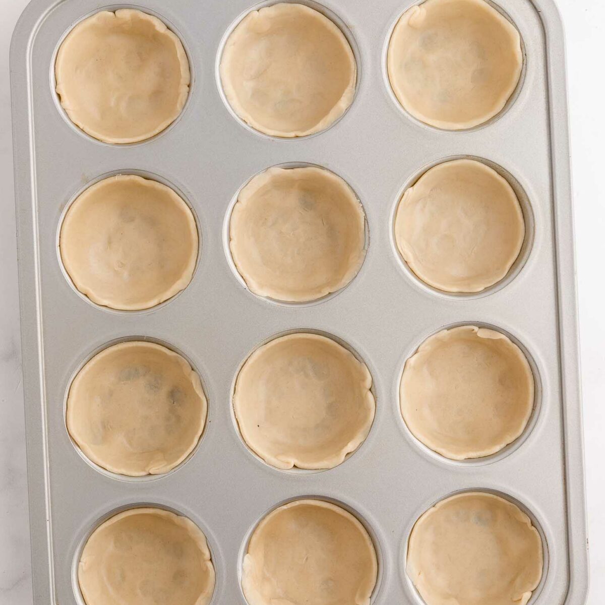 A muffin tin with twelve cavities, each lined with an unbaked, round pie crust, set on a light-colored surface.