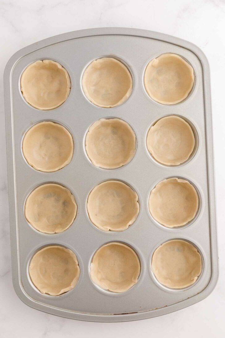 A muffin tin with twelve cavities, each lined with an unbaked, round pie crust, set on a light-colored surface.