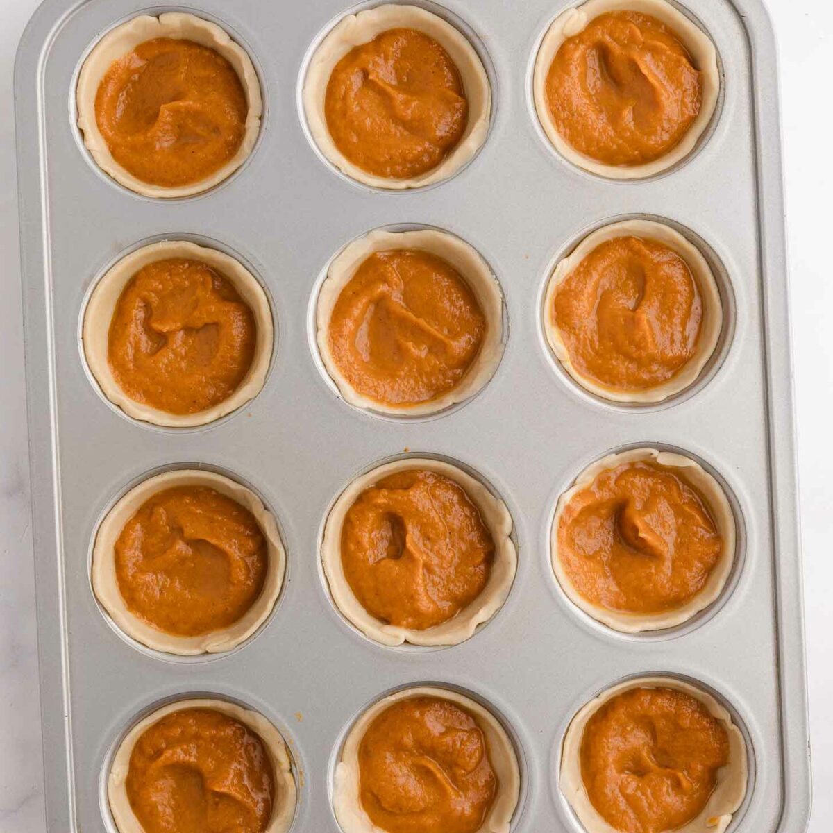 A muffin tin filled with twelve unbaked mini pies, each containing orange-brown pumpkin filling, sits on a white surface.