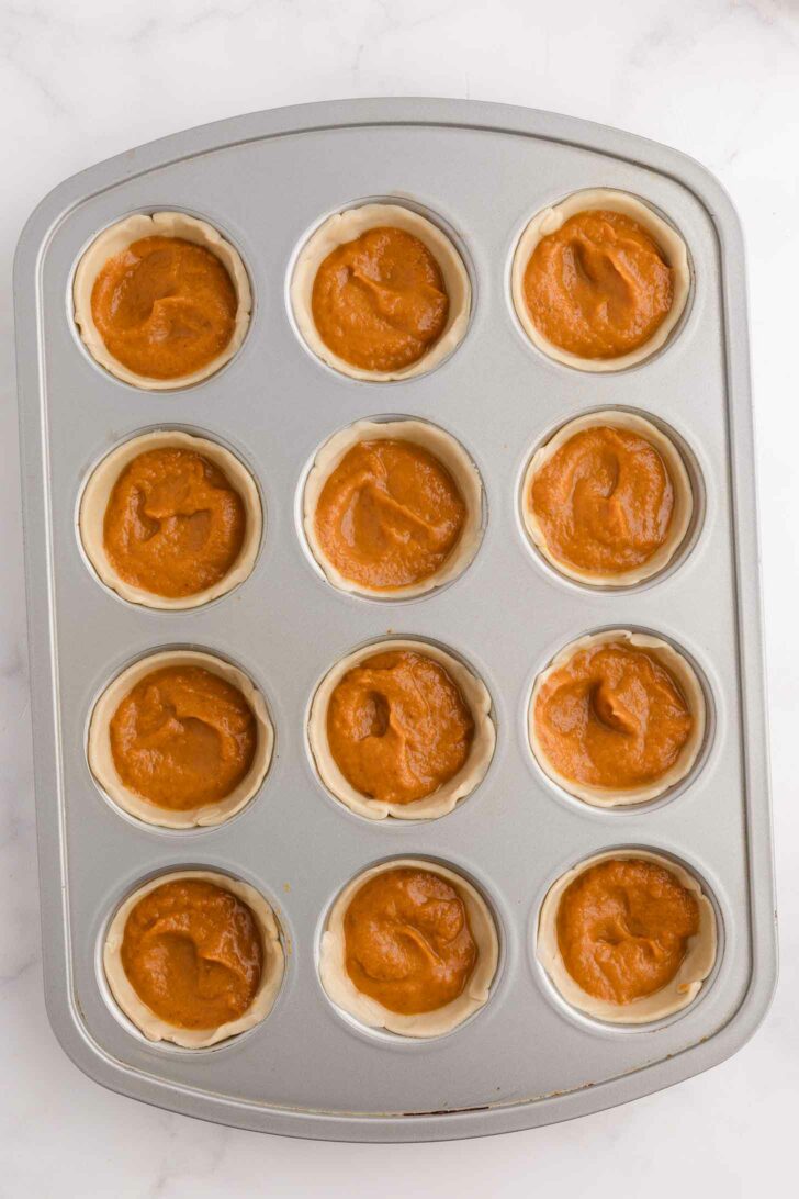A muffin tin filled with twelve unbaked mini pies, each containing orange-brown pumpkin filling, sits on a white surface.