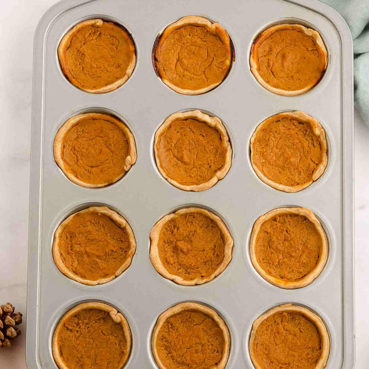 A muffin tin filled with twelve mini pumpkin pies, each with a golden-brown crust and pumpkin filling, sits on a white surface. Nearby are a plate of cinnamon sticks, a green towel, pinecones, and a small pumpkin.