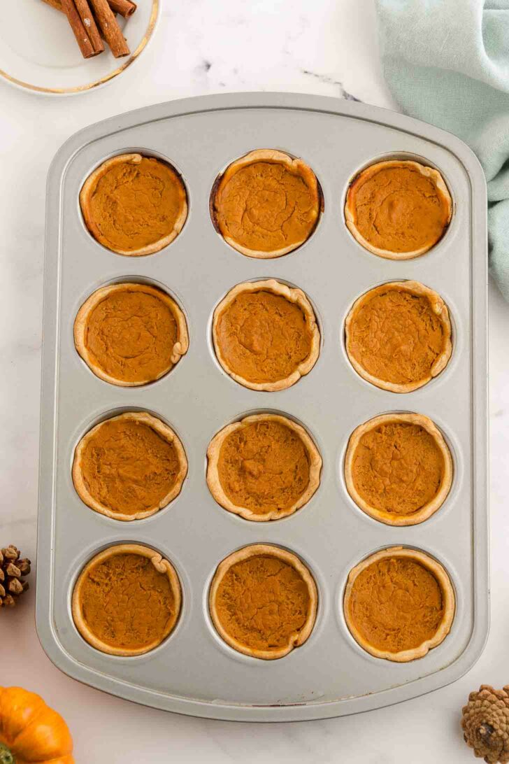 A muffin tin filled with twelve mini pumpkin pies, each with a golden-brown crust and pumpkin filling, sits on a white surface. Nearby are a plate of cinnamon sticks, a green towel, pinecones, and a small pumpkin.