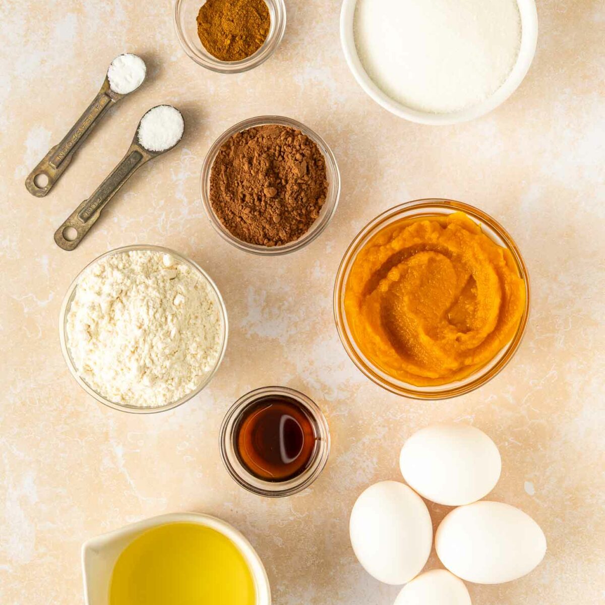 Overhead view of ingredients in small bowls and measuring spoons, including flour, cocoa powder, sugar, pumpkin puree, eggs, vanilla, oil, baking powder, baking soda, and pumpkin pie spice, arranged on a light surface.