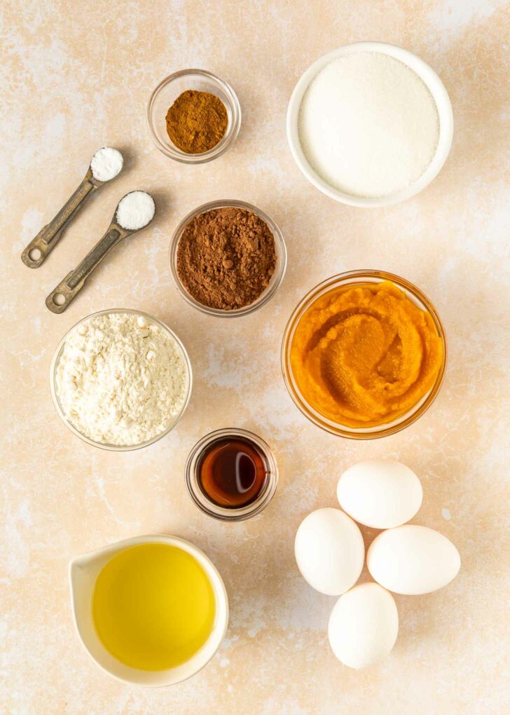 Overhead view of ingredients in small bowls and measuring spoons, including flour, cocoa powder, sugar, pumpkin puree, eggs, vanilla, oil, baking powder, baking soda, and pumpkin pie spice, arranged on a light surface.