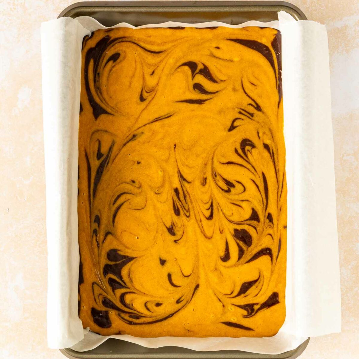 A rectangular tray of pumpkin brownies with a marbled swirl pattern of dark chocolate and orange pumpkin batter, lined with parchment paper.