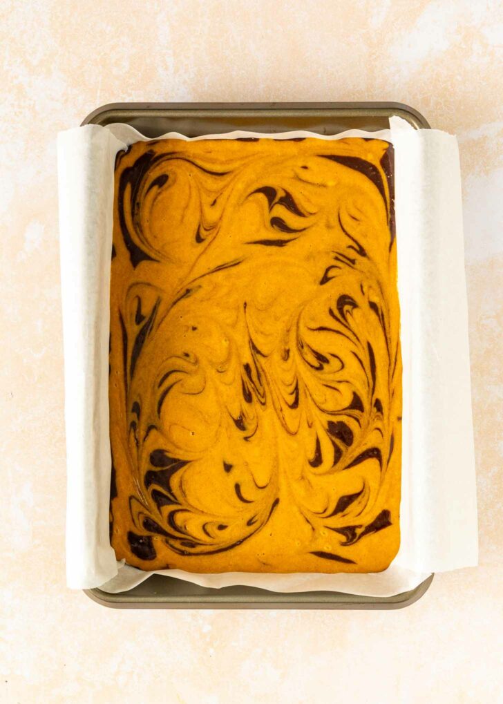 A rectangular tray of pumpkin brownies with a marbled swirl pattern of dark chocolate and orange pumpkin batter, lined with parchment paper.