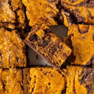 Close-up of pumpkin swirl brownies cut into squares, with one piece slightly raised to show the marbled layers of dark chocolate and orange pumpkin batter.