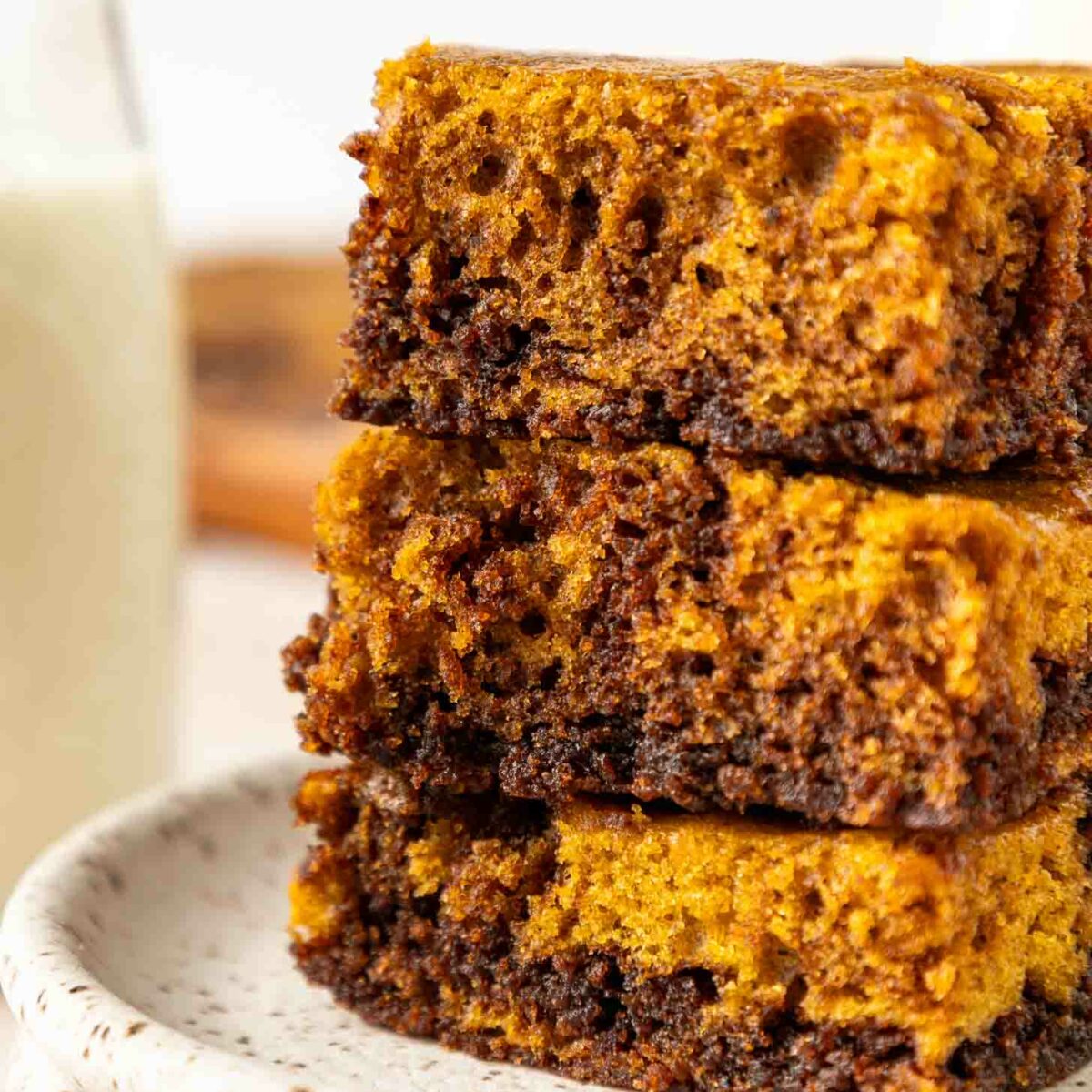 A close-up of three stacked pumpkin chocolate brownies on a speckled ceramic plate, showing their moist, layered texture. A glass of milk is blurred in the background.