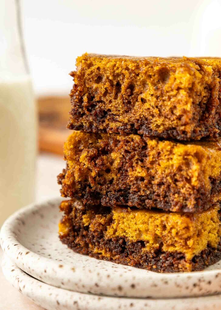 A close-up of three stacked pumpkin chocolate brownies on a speckled ceramic plate, showing their moist, layered texture. A glass of milk is blurred in the background.