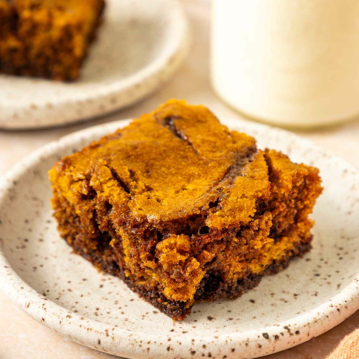 A close-up of a square piece of pumpkin brownie on a speckled white plate, with a blurred background showing another plate of cake and a glass of milk.