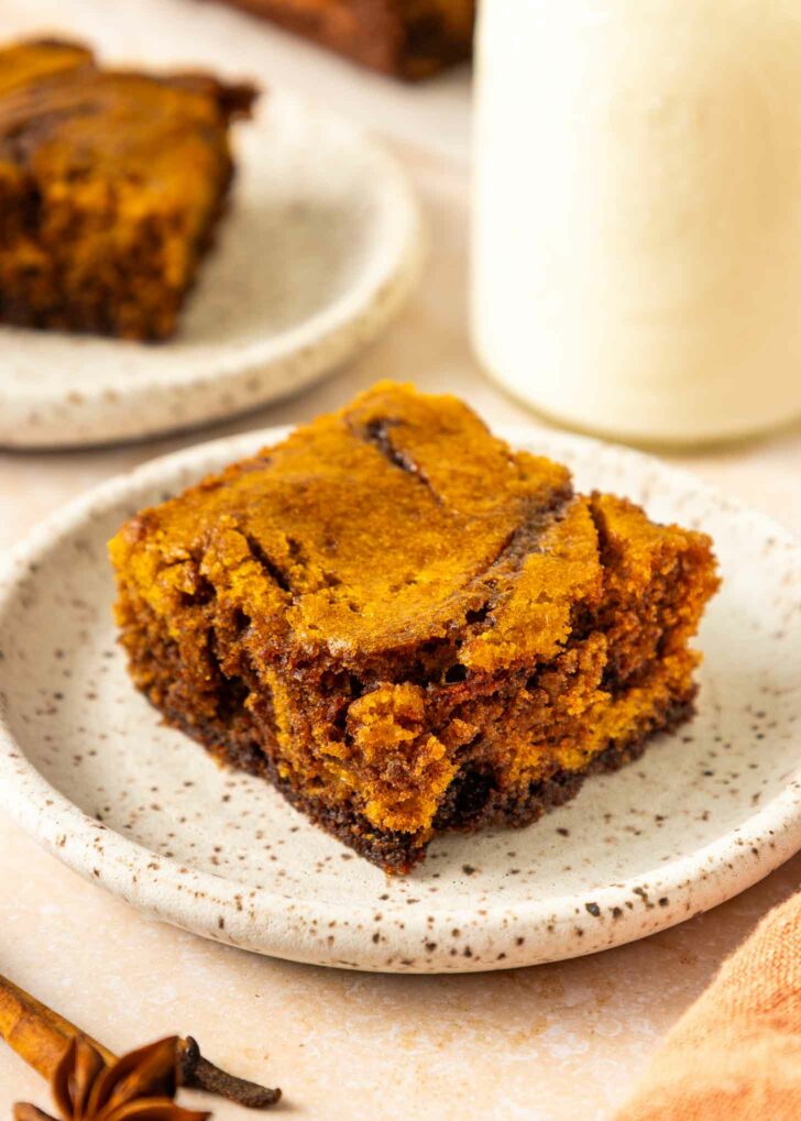 A close-up of a square piece of pumpkin brownie on a speckled white plate, with a blurred background showing another plate of cake and a glass of milk.