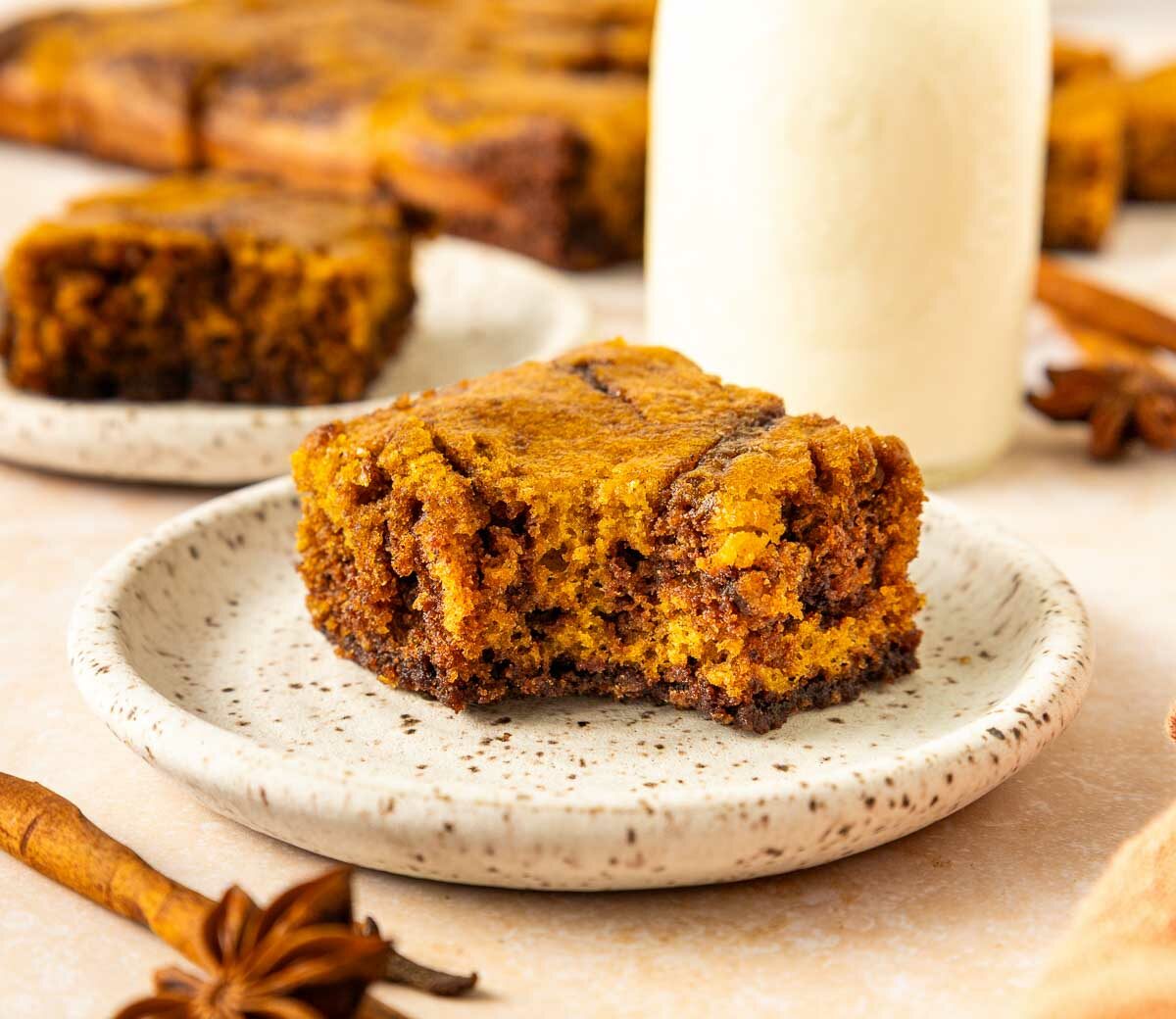 A slice of pumpkin brownie sits on a speckled plate with a blurred background showing another piece, cinnamon sticks, star anise, and a glass bottle of milk.
