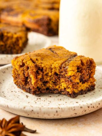 A slice of pumpkin brownie sits on a speckled plate with a blurred background showing another piece, cinnamon sticks, star anise, and a glass bottle of milk.