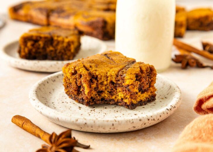 A slice of pumpkin brownie sits on a speckled plate with a blurred background showing another piece, cinnamon sticks, star anise, and a glass bottle of milk.