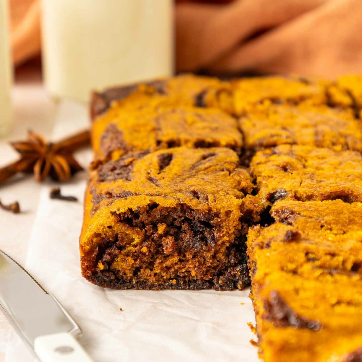 A close-up of a sliced pumpkin brownies on parchment paper, with a knife beside it and glasses of milk and an orange cloth in the background.