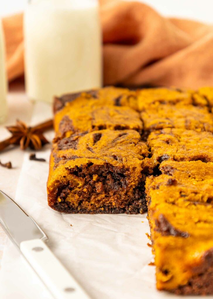 A close-up of a sliced pumpkin brownies on parchment paper, with a knife beside it and glasses of milk and an orange cloth in the background.