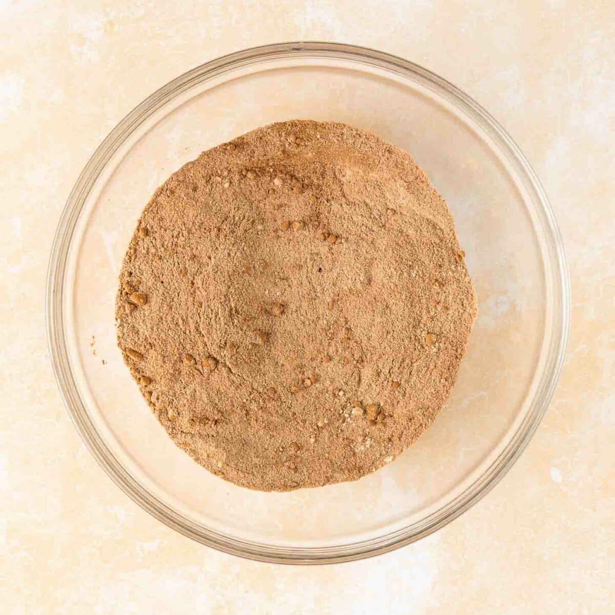 A clear glass bowl filled with a mound of flour, cocoa powder, and other dry ingredients sits on a beige surface.