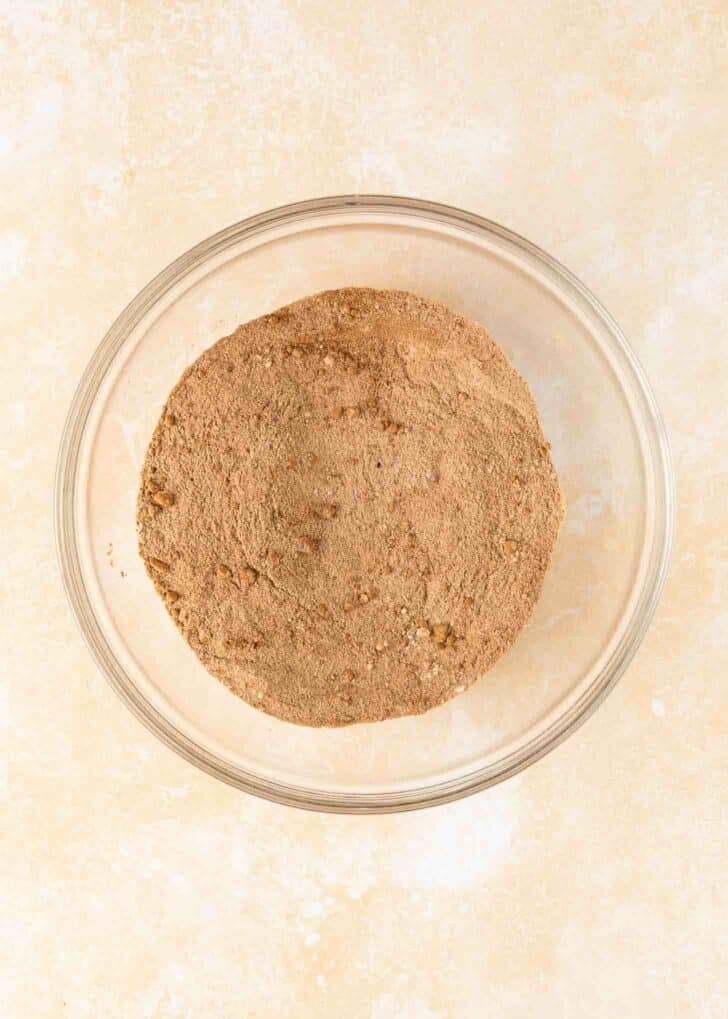 A clear glass bowl filled with a mound of flour, cocoa powder, and other dry ingredients sits on a beige surface.