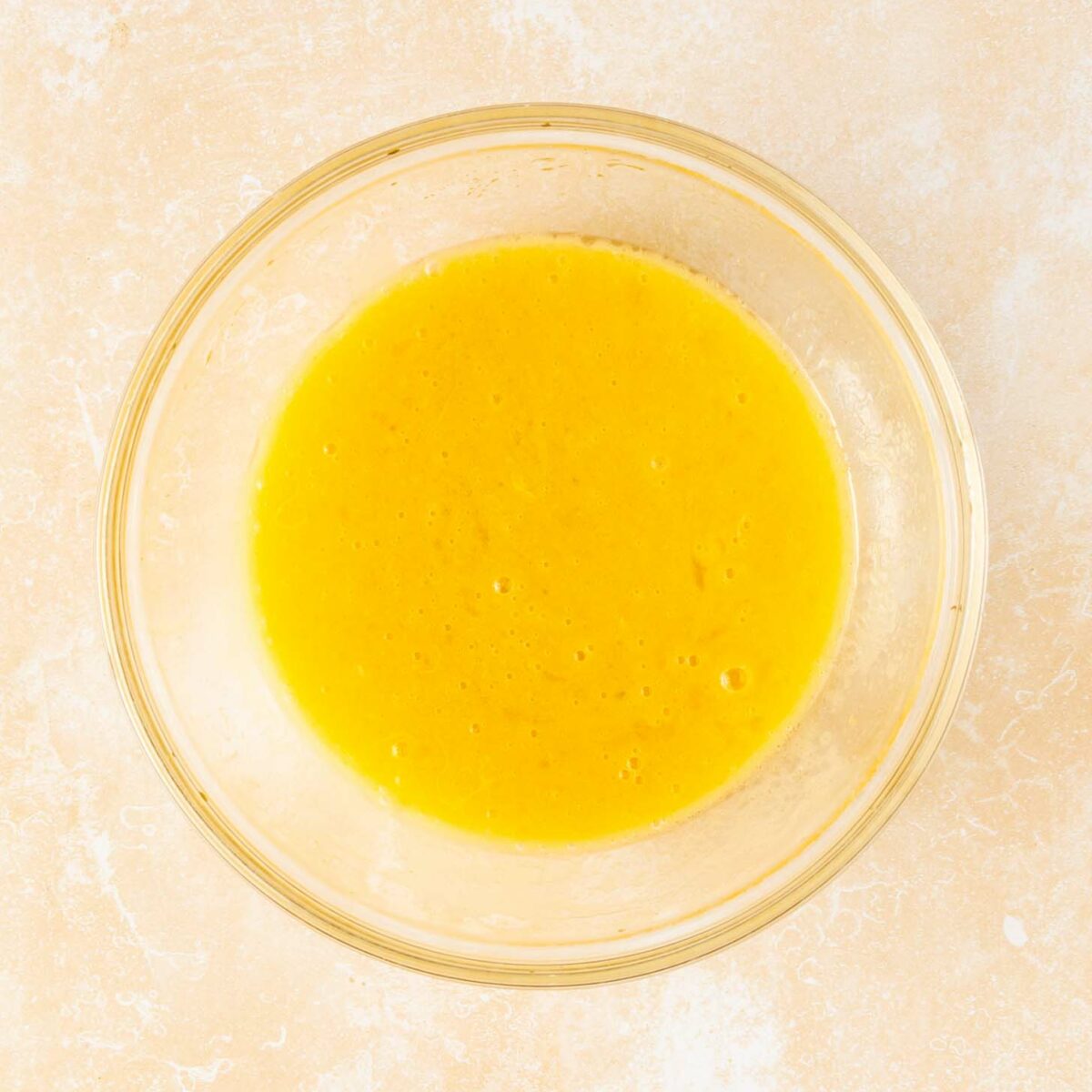 A clear glass bowl filled with beaten yellow eggs sits on a light, textured surface.