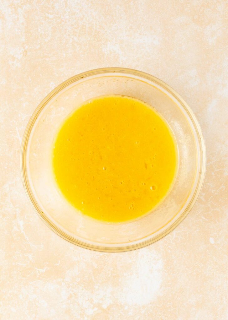 A clear glass bowl filled with beaten yellow eggs sits on a light, textured surface.