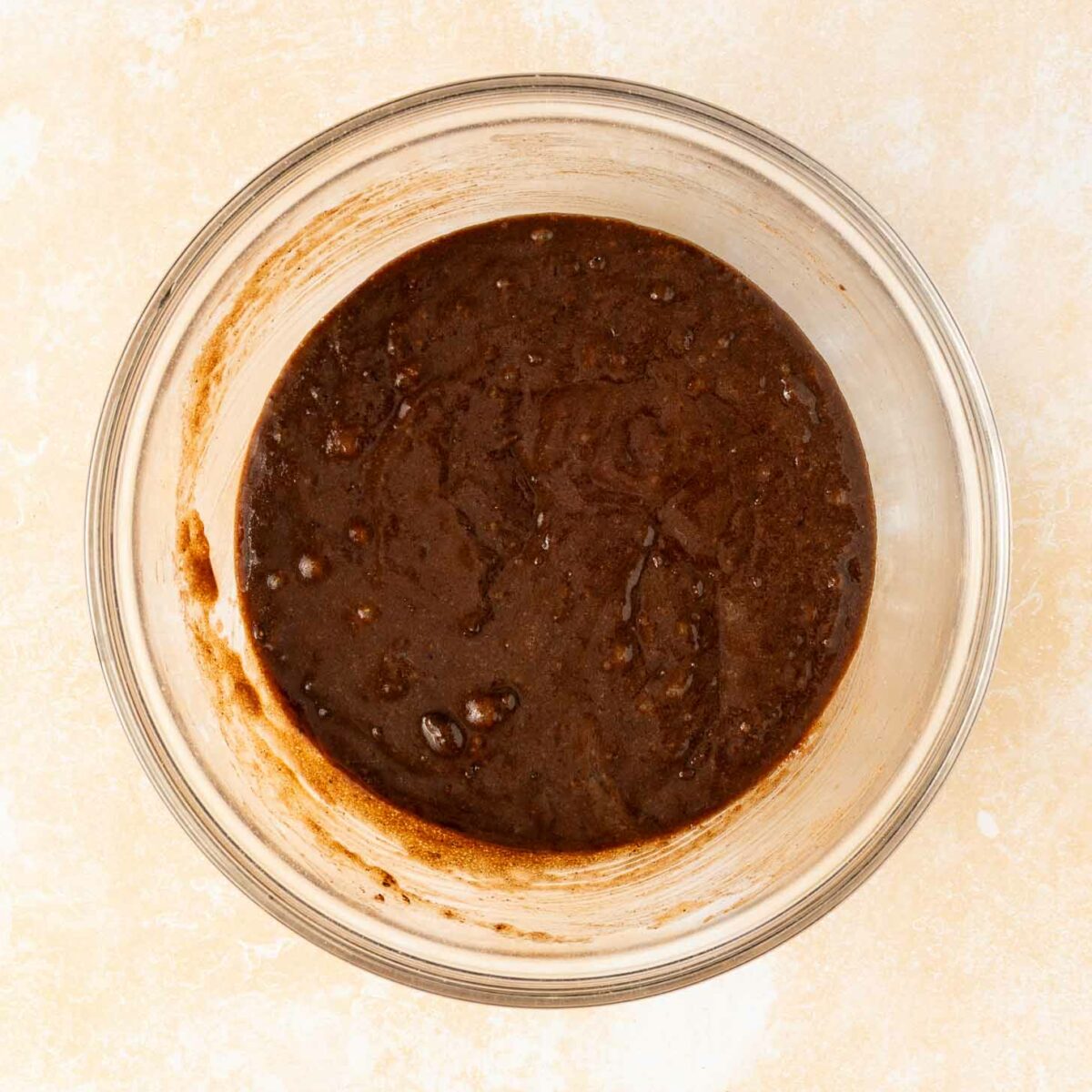 A clear glass bowl containing a thick, dark brownie batter sits on a light, textured surface.