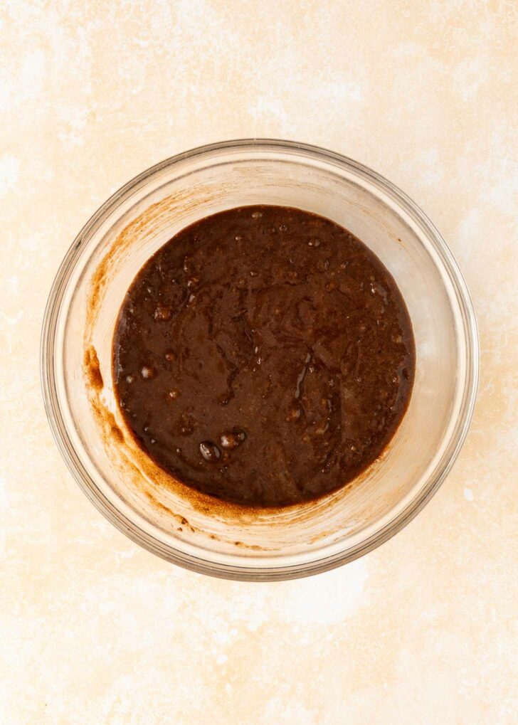 A clear glass bowl containing a thick, dark brownie batter sits on a light, textured surface.