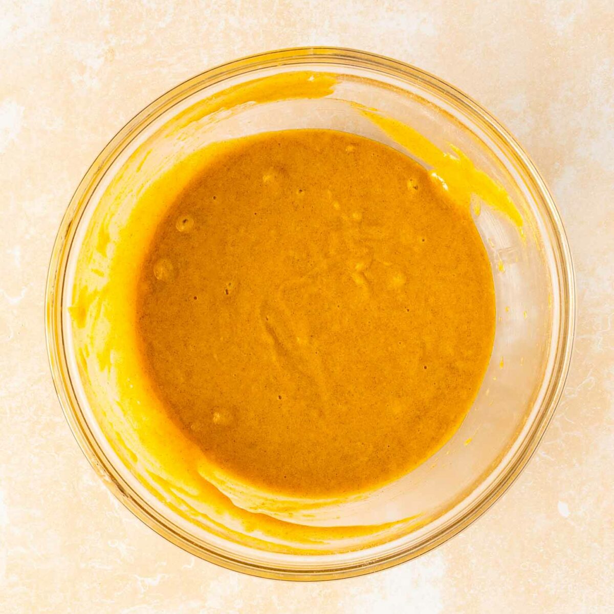 A clear glass bowl filled with smooth, yellowish-orange batter sits on a light, textured surface.