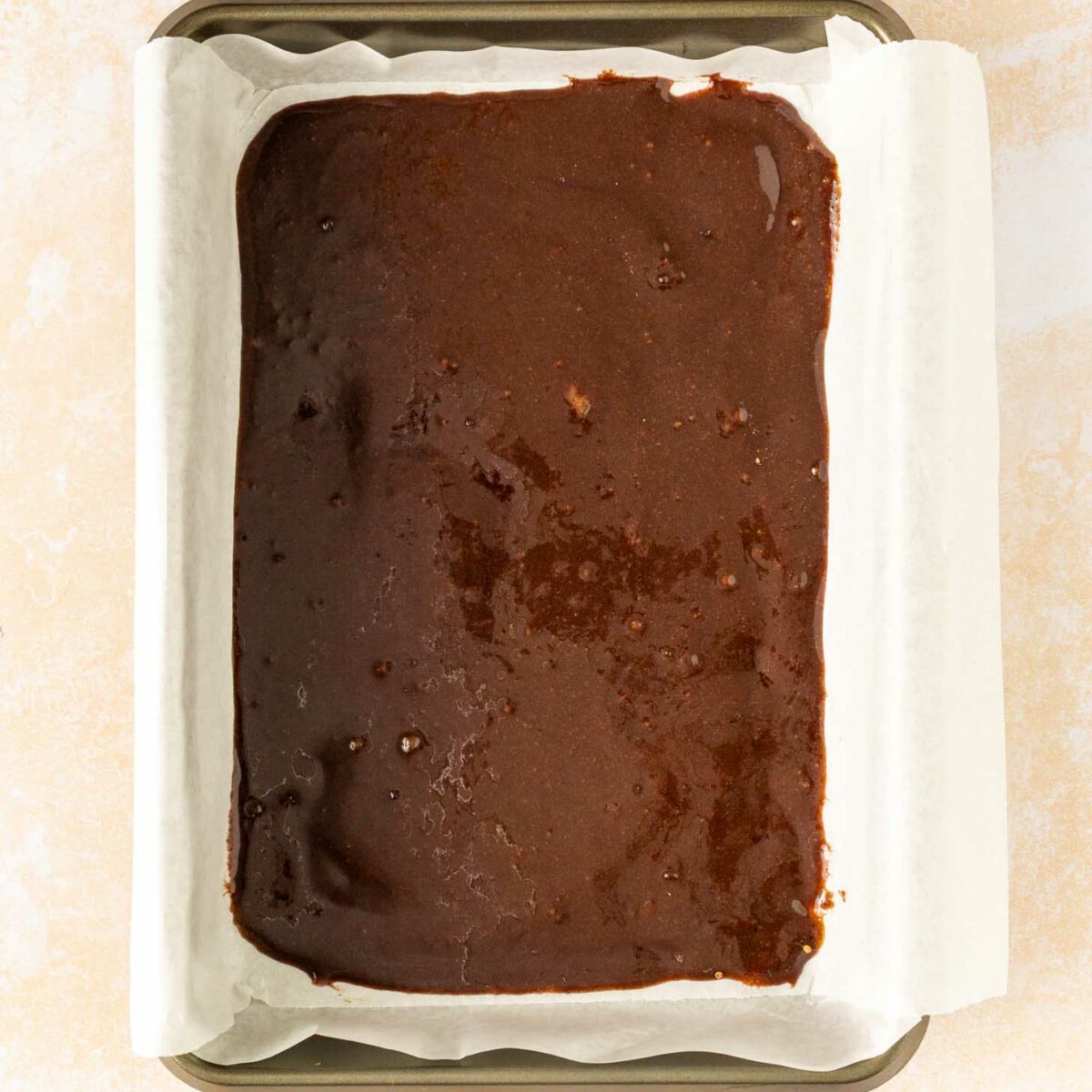 A rectangular baking pan lined with parchment paper is filled with unbaked chocolate brownie batter, ready to go into the oven.