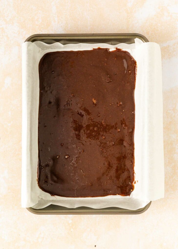 A rectangular baking pan lined with parchment paper is filled with unbaked chocolate brownie batter, ready to go into the oven.
