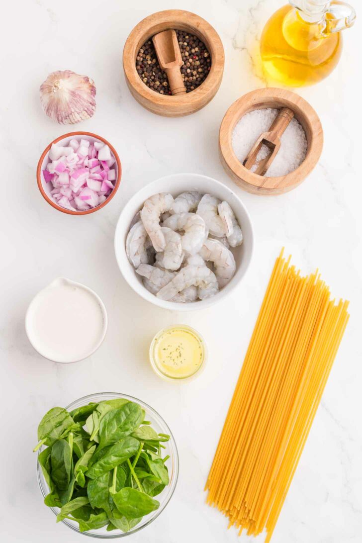 Overhead view of ingredients on a white surface: raw shrimp, dry spaghetti, spinach leaves, chopped red onions, garlic, black pepper, salt, olive oil, half-and-half, and a small bowl of seasoning.