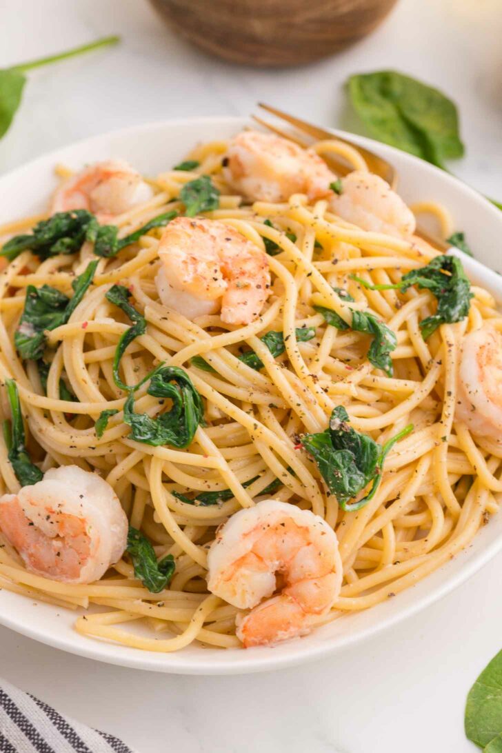 A white bowl filled with spaghetti, sautéed shrimp, and wilted spinach, topped with cracked black pepper, sits on a marble surface.