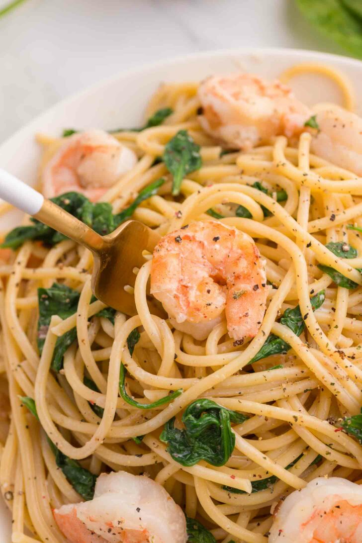 A close-up of a plate of spaghetti pasta mixed with shrimp and fresh spinach, seasoned with black pepper, with a fork twirling some noodles.