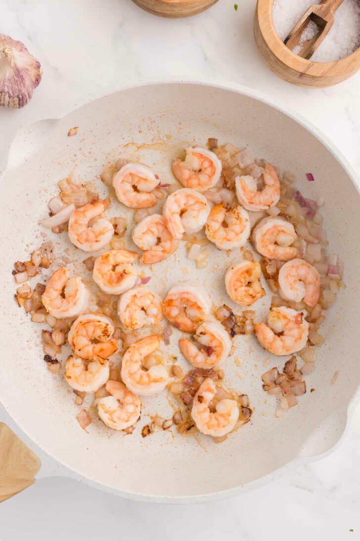Shrimp being sautéed with chopped onions in a white pan, surrounded by bowls of salt and a head of garlic on a white countertop.
