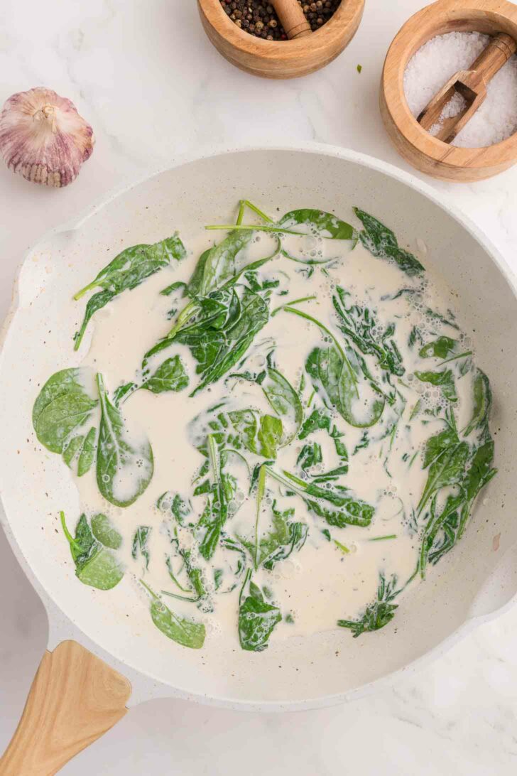 A white skillet filled with fresh spinach leaves cooking in a creamy white sauce, surrounded by a wooden bowl of salt, a bowl of pepper, and a garlic bulb on a white countertop.