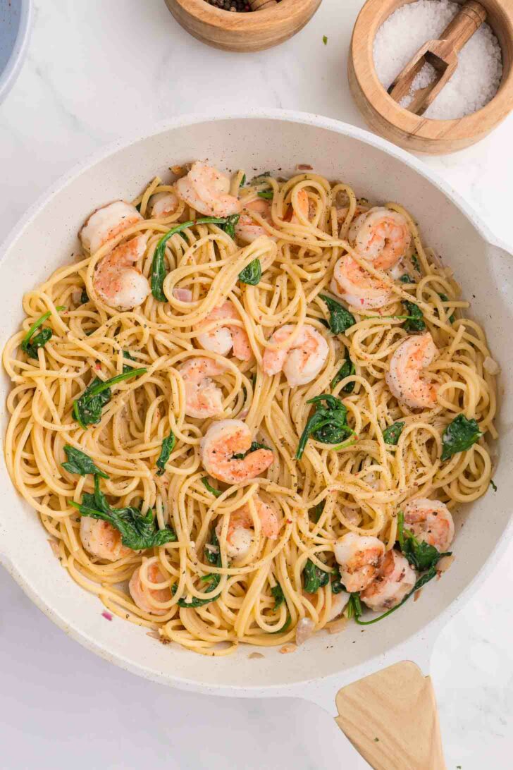 A white skillet filled with spaghetti, sautéed shrimp, and wilted spinach, garnished with herbs and black pepper. Wooden bowls with salt and pepper are nearby on a white countertop.