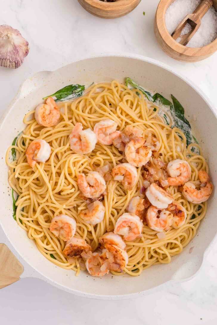 A white skillet filled with cooked spaghetti, shrimp, spinach, and a creamy sauce, placed on a white marble surface with garlic and bowls of salt and pepper nearby.