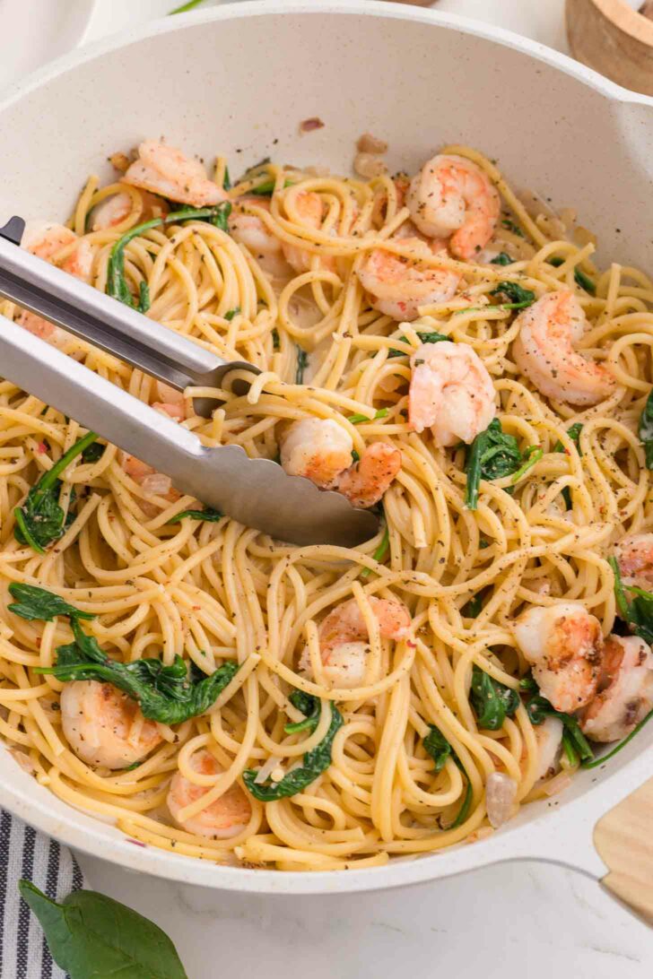 A pan filled with cooked spaghetti, shrimp, and wilted spinach, with metal tongs resting on top, ready to serve.