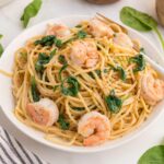 A white bowl filled with spaghetti, shrimp, and spinach on a marble surface, with fresh spinach leaves scattered around and a striped napkin nearby.