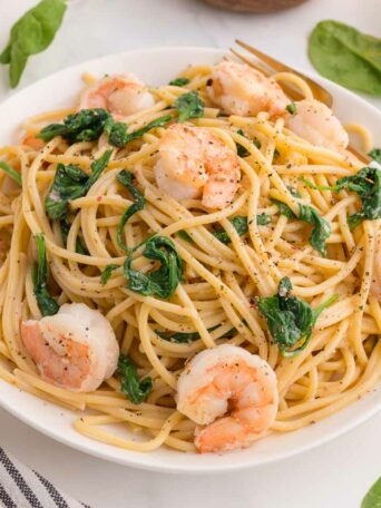 A white bowl filled with spaghetti, shrimp, and spinach on a marble surface, with fresh spinach leaves scattered around and a striped napkin nearby.