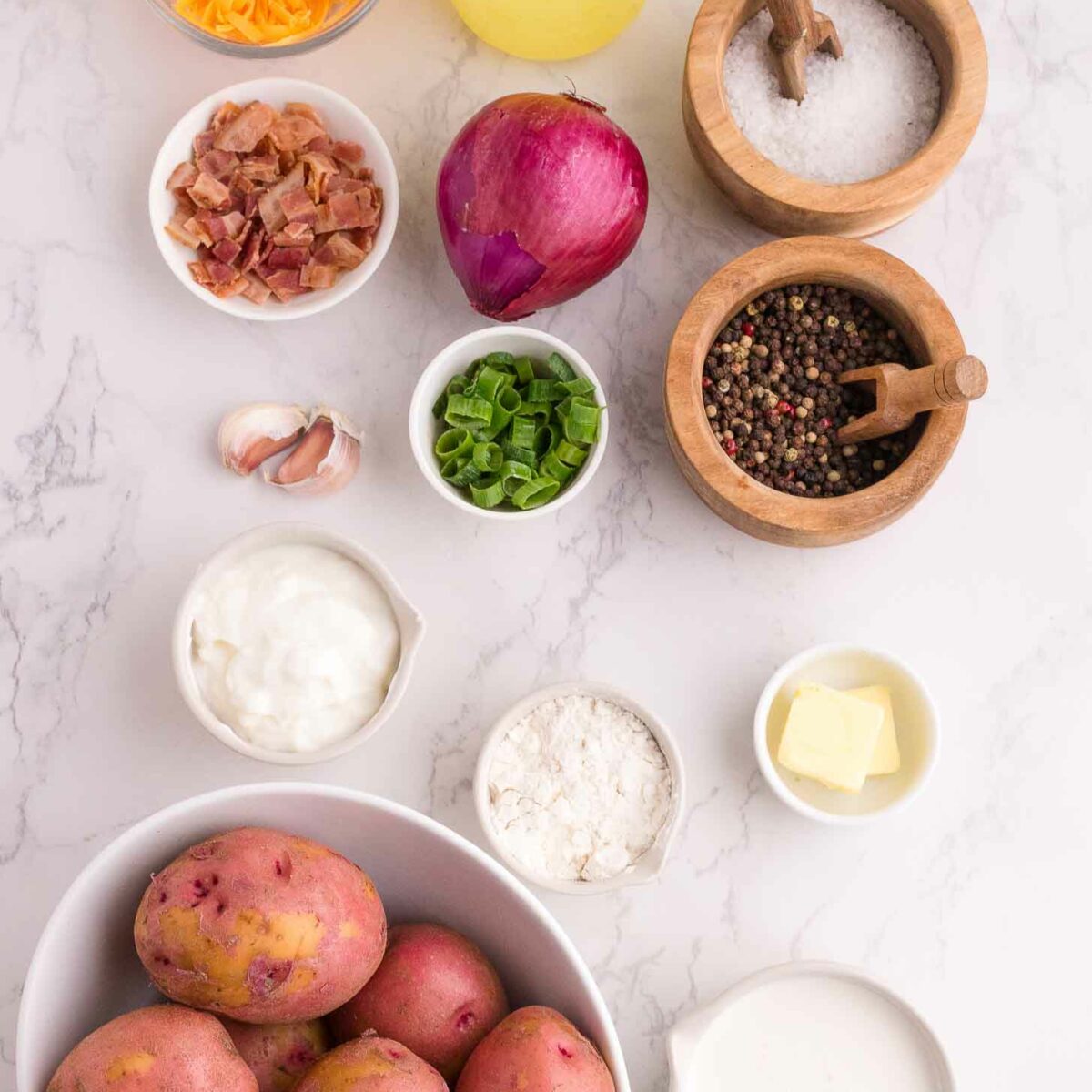 An assortment of loaded potato soup ingredients on a white marble surface, including red potatoes, shredded cheese, chicken broth, bacon, red onion, garlic, green onions, sour cream, flour, butter, milk, salt, and pepper.