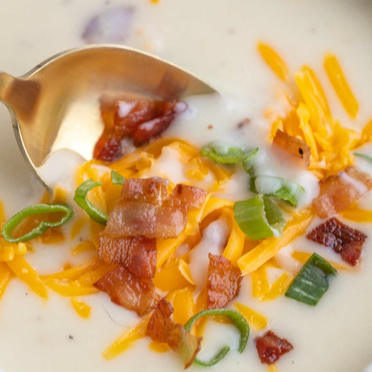 A close-up of creamy potato soup in a white bowl, topped with shredded cheddar cheese, crispy bacon pieces, and sliced green onions. A gold spoon is resting in the soup.