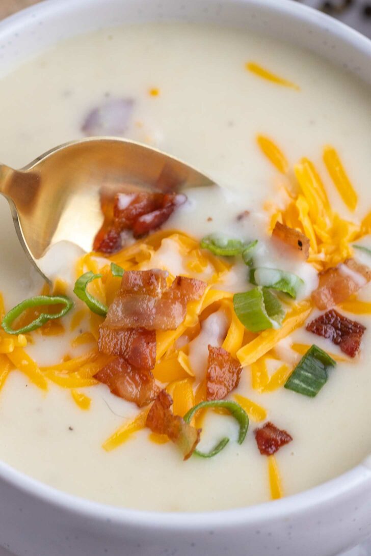 A close-up of creamy potato soup in a white bowl, topped with shredded cheddar cheese, crispy bacon pieces, and sliced green onions. A gold spoon is resting in the soup.