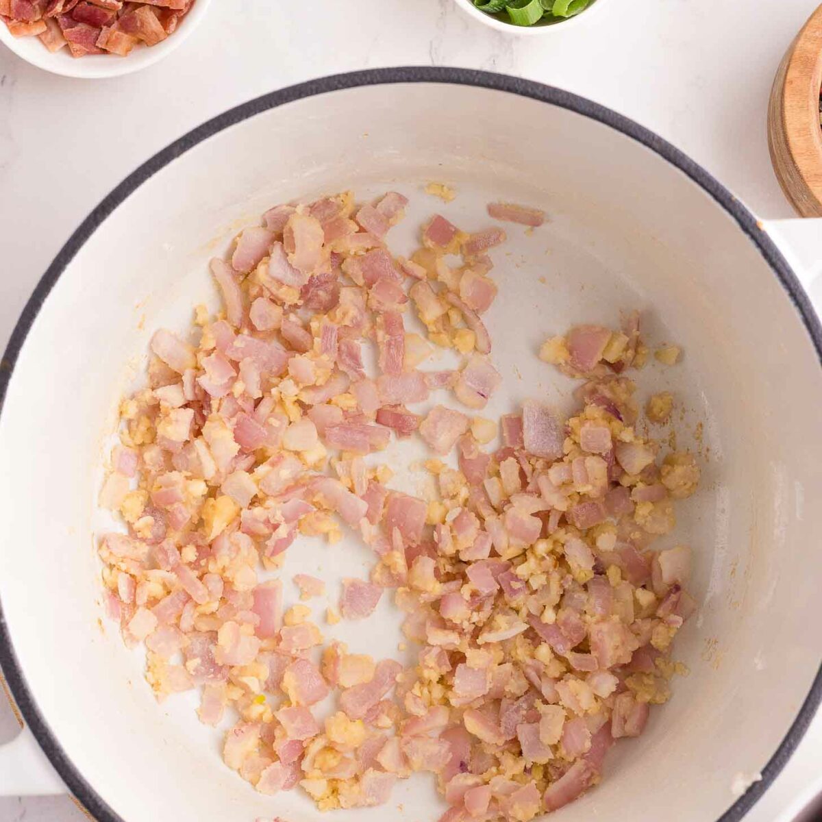 Chopped red onion and garlic cooking in a white pot, viewed from above. Surrounding the pot are small bowls containing cooked bacon pieces and sliced green onions on a marble countertop.