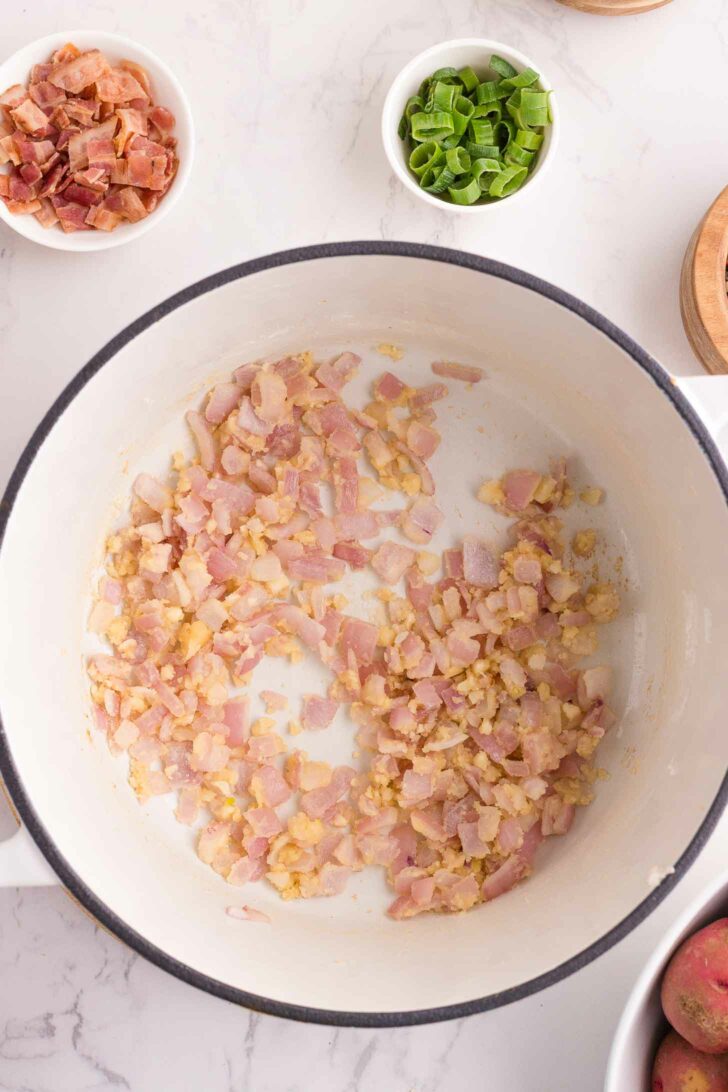 Chopped red onion and garlic cooking in a white pot, viewed from above. Surrounding the pot are small bowls containing cooked bacon pieces and sliced green onions on a marble countertop.