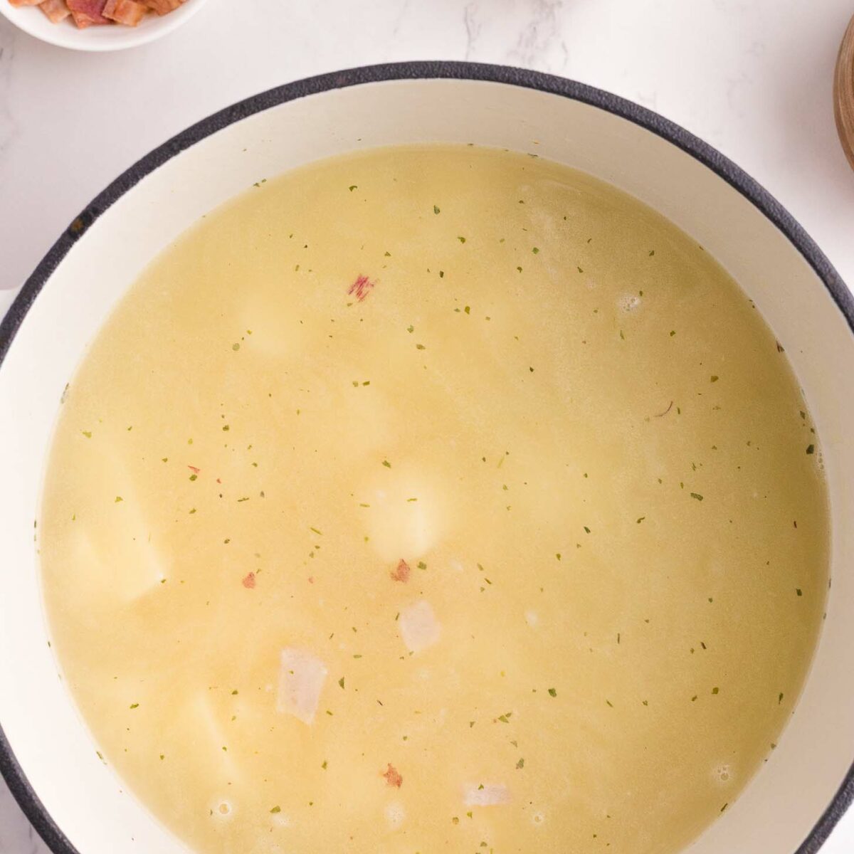 A pot of light-colored soup with visible potato chunks sits on a white surface. Nearby, small bowls hold chopped bacon and sliced green onions.