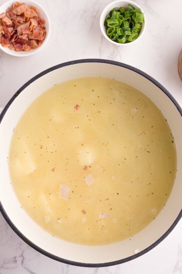 A pot of light-colored soup with visible potato chunks sits on a white surface. Nearby, small bowls hold chopped bacon and sliced green onions.