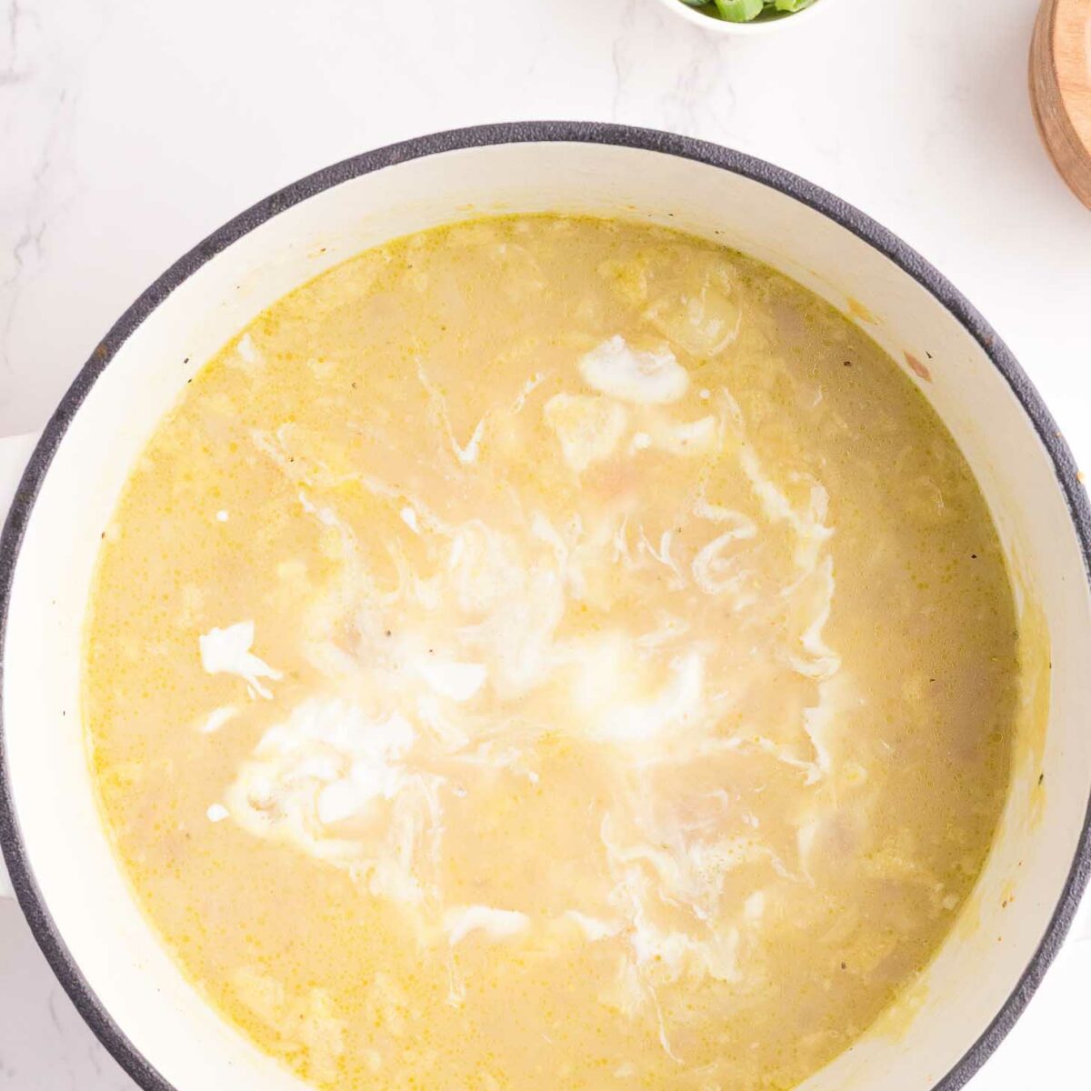 A pot of creamy soup with white swirls on top sits on a light marble surface. A small bowl of chopped green onions is nearby for garnish.