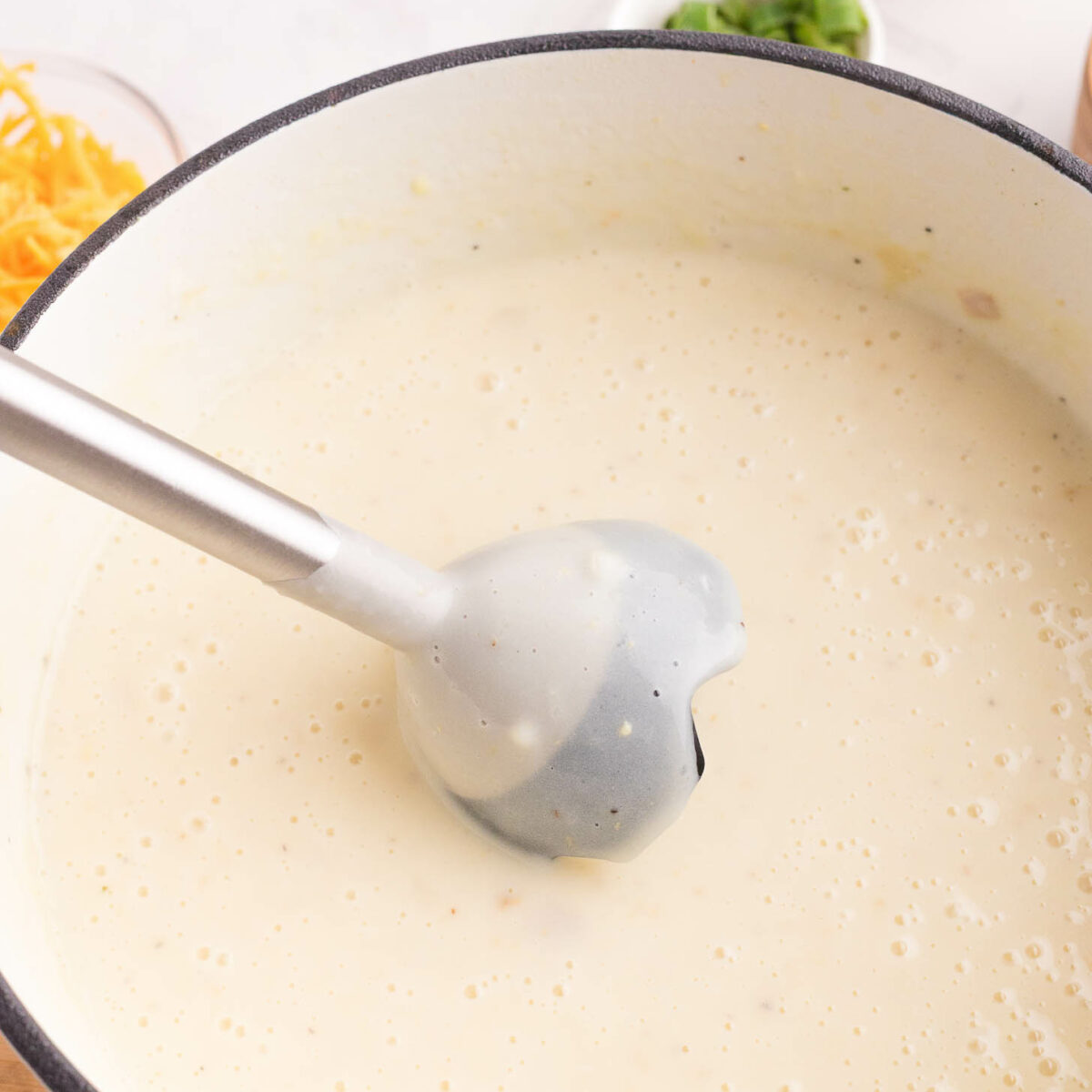 A large pot filled with creamy, light-colored soup, with an immersion blender partially submerged. Ingredients like shredded cheese and chopped green onions are visible in small bowls nearby.